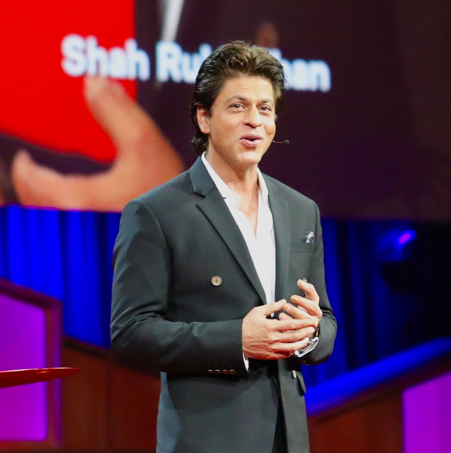 Shah Rukh Khan speaking on stage in a dark double-breasted blazer and white open-collar shirt